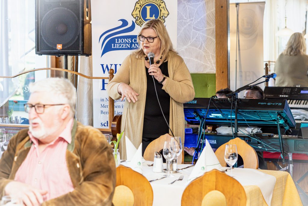 Eine Frau spricht bei einer Veranstaltung des Lions Club Liezen Anasia ins Mikrofon. Im Hintergrund stehen Keyboard und Lautsprecher der Musiktechnik, während Gäste an festlich gedeckten Tischen sitzen und der Ansprache zuhören.