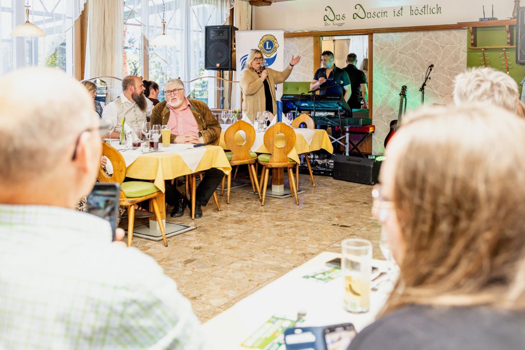 Eine Frau spricht bei einer Veranstaltung des Lions Club Liezen Anasia vor den Gästen im Restaurant. Die Besucher sitzen an gedeckten Tischen und verfolgen die Ansprache, während im Hintergrund Musik- und Veranstaltungstechnik aufgebaut ist.