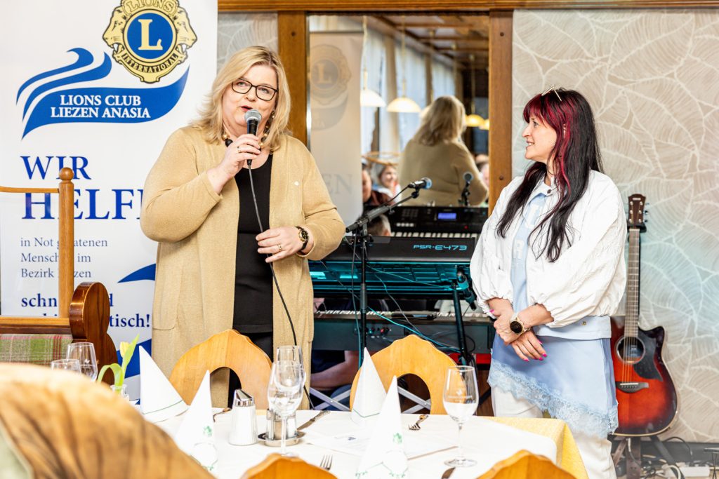 Eine Frau spricht bei einer Veranstaltung des Lions Club Liezen Anasia ins Mikrofon, während eine Teilnehmerin der Modenschau aufmerksam daneben steht. Im Hintergrund sind Musikinstrumente und Veranstaltungstechnik zu sehen, davor gedeckte Tische mit Gästen.