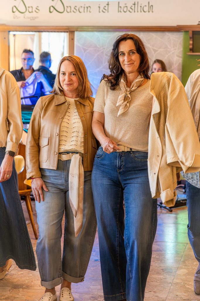 Zwei Frauen präsentieren bei einer Modenschau lässige Frühlingslooks in Beige- und Blautönen. Die eine trägt eine beige Lederjacke mit hellem Stricktop und Jeans, die andere kombiniert ein feines Strickshirt mit dunklen Wide-Leg-Jeans und einem locker über die Schulter gelegten Cardigan.