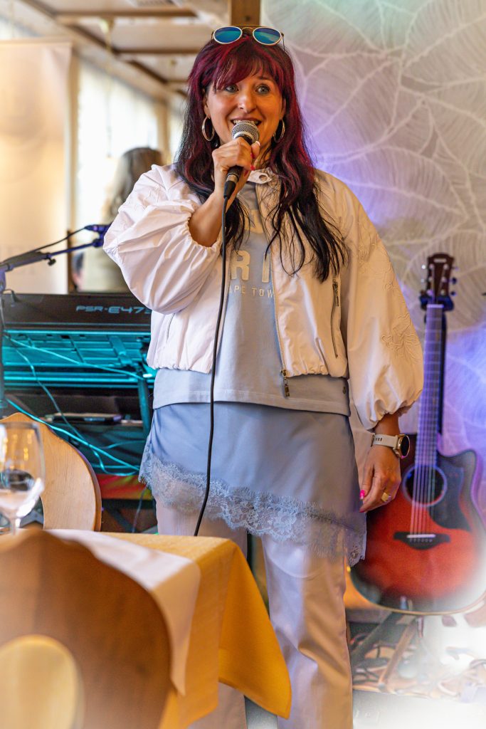 Eine Frau mit roten Haaren spricht bei einer Veranstaltung in einem Restaurant in ein Mikrofon. Sie trägt ein hellblaues Outfit mit weißer Jacke und steht vor Musik-Equipment und einer Gitarre, während die warme Beleuchtung für eine gemütliche Atmosphäre sorgt.