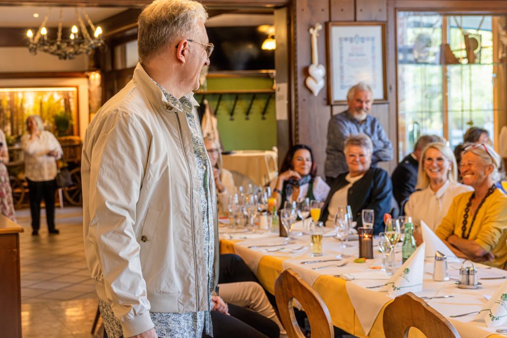 Ein Mann in heller Jacke präsentiert bei einer Modeschau ein elegantes Freizeitoutfit in einem Restaurant. Die Gäste sitzen an festlich gedeckten Tischen, lachen und beobachten aufmerksam die Veranstaltung in der warmen, gemütlichen Atmosphäre.