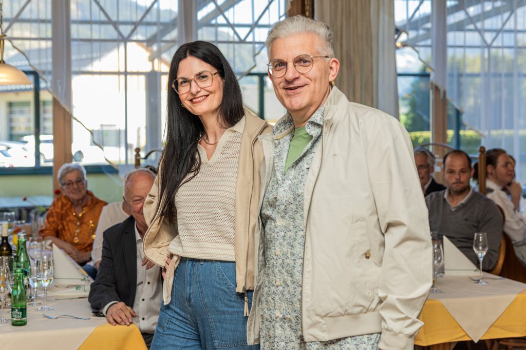 Ein Mann und eine Frau posieren gemeinsam bei einer Modeschau in einem Restaurant. Beide tragen helle Jacken und elegante Freizeitmode mit Jeans und lächeln in die Kamera, während Gäste an den Tischen die Veranstaltung verfolgen.