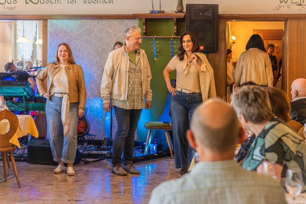 Drei Personen präsentieren bei einer Modeschau moderne Casual-Outfits mit Jeans und hellen Naturtönen in einem Restaurant. Zwei Frauen und ein Mann posieren selbstbewusst vor den Gästen, während im Hintergrund Musik-Equipment und warme Beleuchtung für eine stilvolle Atmosphäre sorgen.