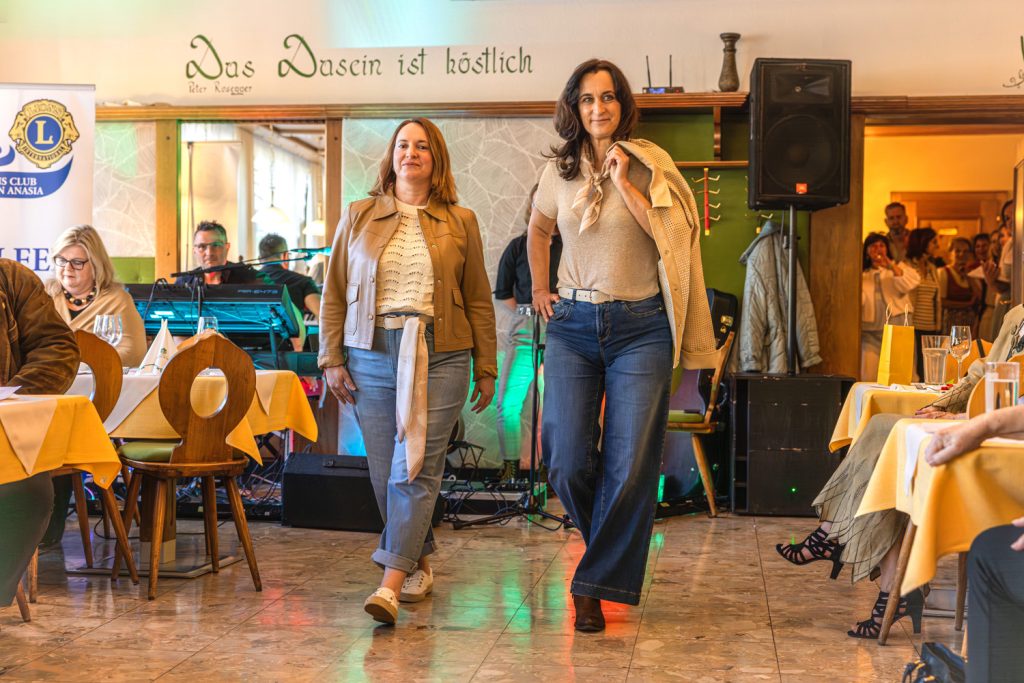 Zwei Frauen präsentieren bei einer Modeschau lässig-elegante Outfits mit Jeans in einem Restaurant. Eine trägt eine braune Lederjacke über einem hellen Oberteil, die andere kombiniert eine beige Strickoptik mit weiter Jeans und einem Tuch über der Schulter. Gäste sitzen an den Tischen und verfolgen die Veranstaltung, während im Hintergrund Musik-Equipment und stimmungsvolle Beleuchtung zu sehen sind.