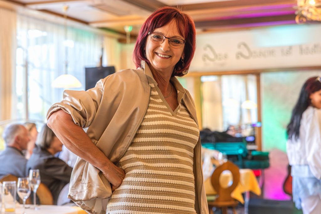 Eine lächelnde Frau mit roten Haaren und Brille posiert bei einer Modeschau in einem Restaurant. Sie trägt eine beige Jacke über einem gestreiften Oberteil und steht selbstbewusst mit der Hand in der Hüfte, während im Hintergrund Gäste und bunte Bühnenbeleuchtung zu sehen sind.