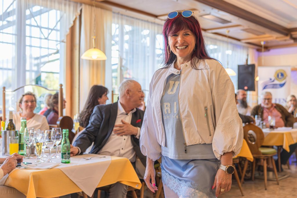 Eine lächelnde Frau mit roten Haaren präsentiert bei einer Modeschau ein hellblaues Outfit mit weißer Jacke in einem gemütlichen Restaurant. Im Hintergrund sitzen Gäste an gedeckten Tischen und beobachten die Veranstaltung, während warmes Licht durch die großen Fenster fällt.