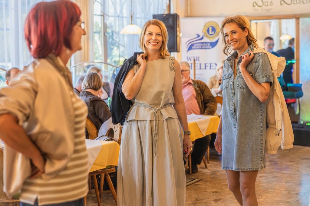 Drei Frauen präsentieren bei einer Modeschau elegante Frühlingsoutfits in einem gemütlichen Restaurant. Zwei Frauen stehen lächelnd im Mittelpunkt – eine in einem hellen Kleid mit schwarzer Jacke über der Schulter, die andere in einem kurzen Jeanskleid mit heller Jacke. Im Vordergrund ist eine weitere Frau leicht unscharf zu sehen, während Gäste an den Tischen das Event verfolgen.