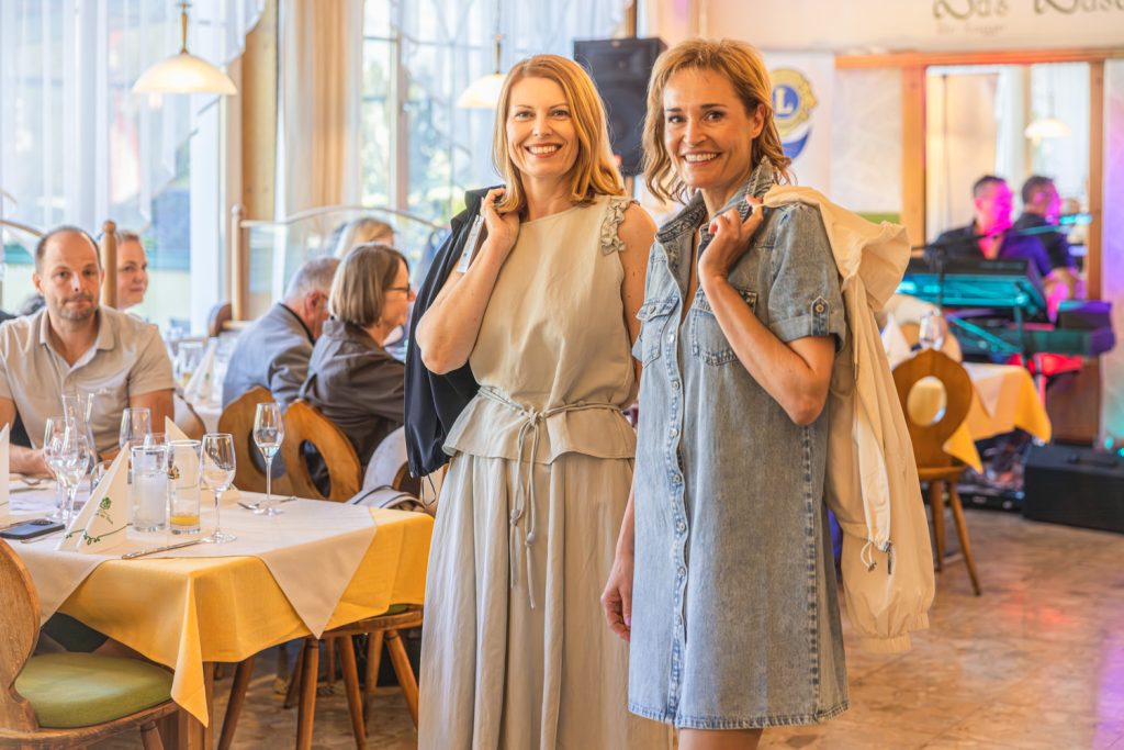 Zwei lächelnde Frauen posieren bei einer Modeschau in einem Restaurant. Die Frau links trägt ein helles, fließendes Kleid mit schwarzer Jacke über der Schulter, die Frau rechts ein kurzes Jeanskleid mit heller Jacke. Im Hintergrund sitzen Gäste an gedeckten Tischen, während Musik und farbige Beleuchtung für eine festliche Atmosphäre sorgen.