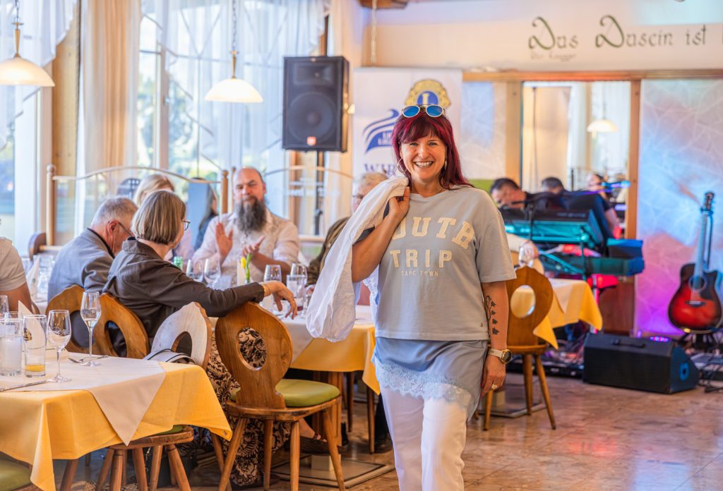 Eine lächelnde Frau mit rot-schwarzen Haaren präsentiert bei einer Modeschau ein hellblaues Freizeit-Outfit mit weißer Hose. Über der Schulter trägt sie eine weiße Jacke, auf dem Kopf eine Sonnenbrille. Im Hintergrund sitzen Gäste an gedeckten Tischen, während Musik-Equipment und eine Gitarre die gemütliche Atmosphäre der Veranstaltung unterstreichen.