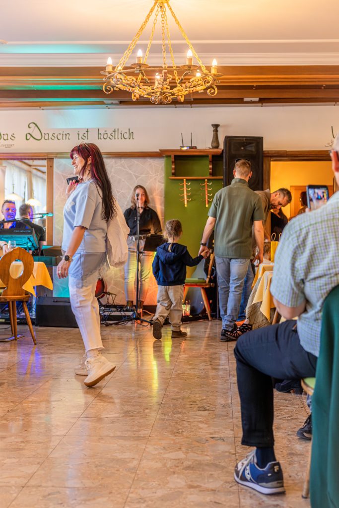 Eine Frau mit rot-schwarzen Haaren läuft bei einer Modeschau durch ein Restaurant und trägt ein hellblaues Outfit mit weißen Sneakers. Im Hintergrund hält ein Mann die Hand eines kleinen Kindes, während weitere Gäste und Teilnehmer der Veranstaltung im Raum verteilt sind. Die Szene ist von warmem Licht, einem Kronleuchter und bunter Bühnenbeleuchtung geprägt, während Zuschauer an den Tischen sitzen und die Modenschau verfolgen.
