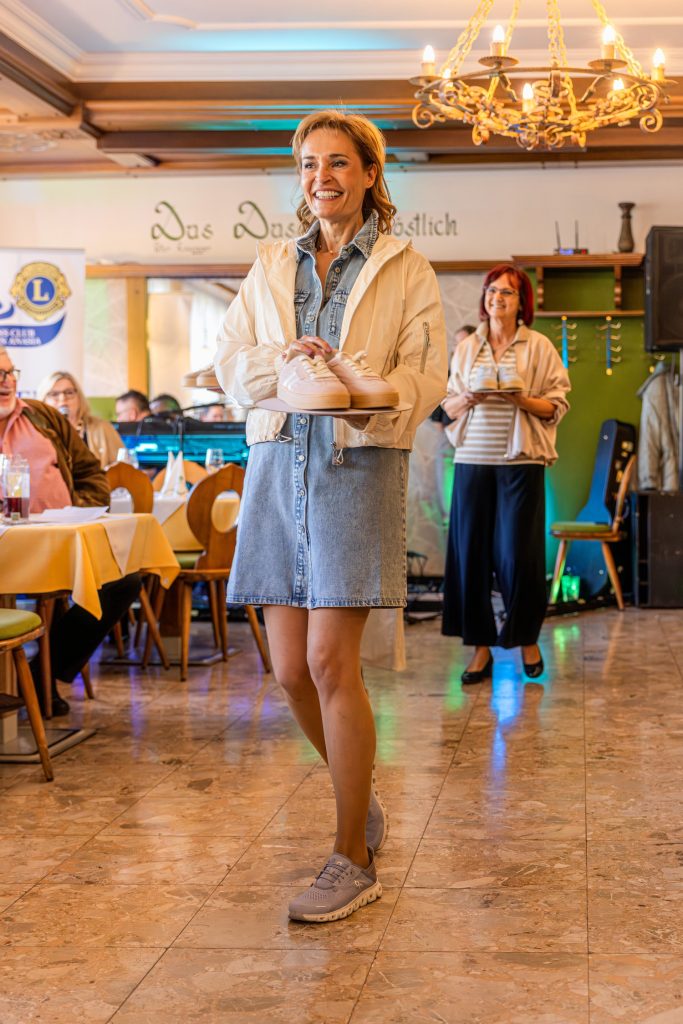Eine lächelnde Frau präsentiert bei einer Modeschau ein Paar rosafarbene Sneakers auf einem weißen Teller. Sie trägt ein kurzes Jeanskleid mit heller Jacke und sportlichen Schuhen. Im Hintergrund folgt eine weitere Frau mit roten Haaren, die ebenfalls Sneakers präsentiert. Die Veranstaltung findet in einem gemütlichen Restaurant mit Kronleuchter, gedeckten Tischen und farbiger Beleuchtung statt.