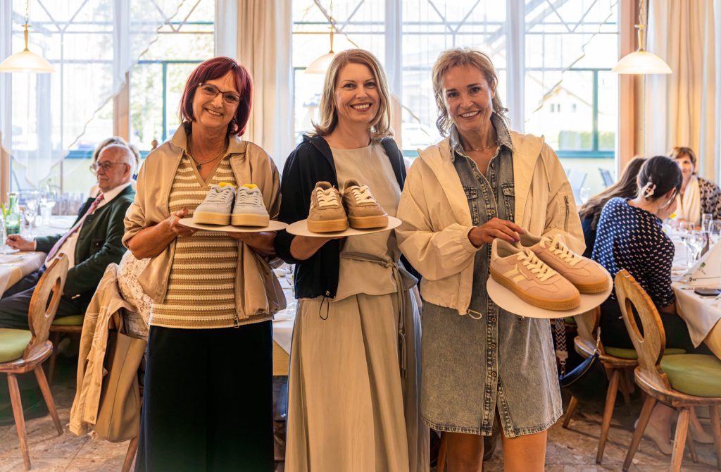 Drei Frauen präsentieren bei einer Modeschau jeweils ein Paar Sneakers auf weißen Tellern. Die Frau links trägt eine beige Jacke mit gestreiftem Oberteil und hält helle Sneaker, die Frau in der Mitte kombiniert ein helles Kleid mit dunkler Jacke und zeigt beigefarbene Schuhe, während die Frau rechts ein Jeanskleid mit weißer Jacke trägt und rosafarbene Sneakers präsentiert. Im Hintergrund sitzen Gäste an gedeckten Tischen vor großen Fenstern und beobachten die Veranstaltung in heller, freundlicher Atmosphäre.