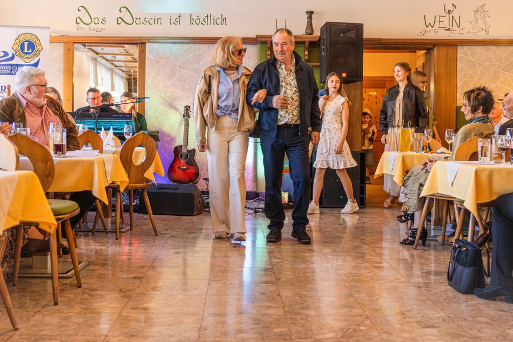 Ein älteres Paar präsentiert Mode bei einer Laufstegshow in einem Restaurant. Die Frau trägt eine Sonnenbrille, eine hellblaue Bluse mit beiger Jacke und helle Hosen, während der Mann ein gemustertes Hemd mit dunkler Jacke und Hose kombiniert. Im Hintergrund warten weitere Teilnehmerinnen der Modenschau, darunter ein Mädchen in einem weißen Sommerkleid und eine junge Frau mit Lederjacke. Gäste sitzen links und rechts an den Tischen und beobachten die Show in gemütlicher Atmosphäre mit Musik-Equipment und bunten Lichteffekten im Hintergrund.