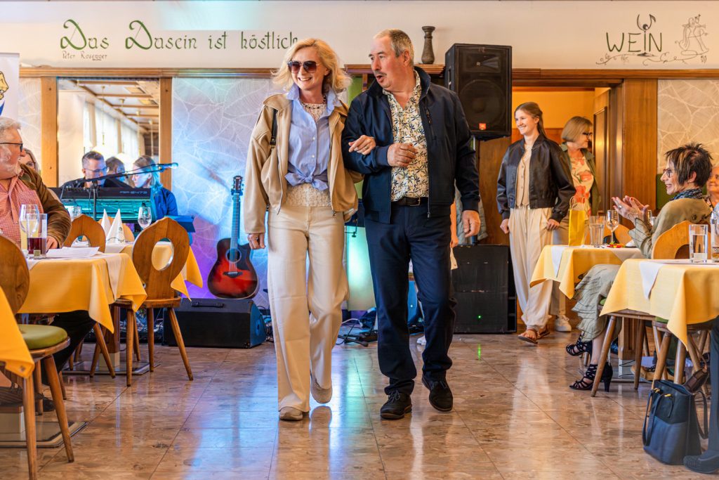 Ein älteres Paar läuft gut gelaunt gemeinsam über den Laufsteg einer Modenschau in einem gemütlichen Restaurant. Die Frau trägt Sonnenbrille, hellblaue Bluse, beige Jacke und helle Hose, während der Mann ein gemustertes Hemd mit dunkler Jacke und Hose kombiniert. Im Hintergrund warten weitere Teilnehmerinnen auf ihren Auftritt, während Gäste an den Tischen sitzen und applaudieren. Bunte Bühnenlichter und warme Holzvertäfelungen schaffen eine festliche Atmosphäre.