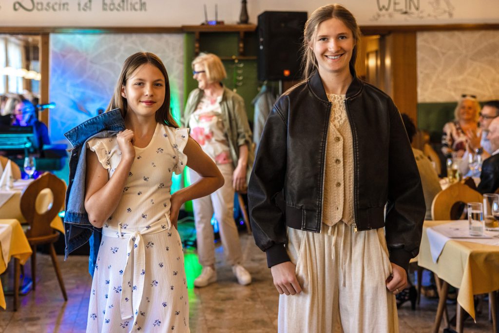 Zwei junge Mädchen präsentieren bei einer Modenschau stilvolle Outfits in einem gemütlichen Veranstaltungsraum. Das Mädchen links trägt ein helles Sommerkleid mit kleinen blauen Mustern und hat eine Jeansjacke locker über die Schulter gelegt. Das Mädchen rechts kombiniert eine helle Hose und Weste mit einer schwarzen Lederjacke. Im Hintergrund beobachten Gäste die Show, während bunte Bühnenlichter für eine festliche Stimmung sorgen.