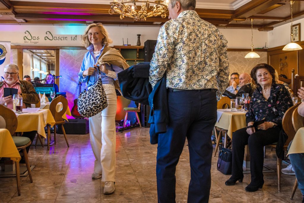 Bei einer Modenschau in einem gemütlichen Restaurant begegnen sich ein älterer Mann und eine blonde Frau auf dem Laufsteg. Die Frau trägt ein frisches Frühlingsoutfit mit hellblauer Bluse, weißer Hose und einer großen Tasche mit Leopardenmuster, während der Mann ein gemustertes Hemd und dunkle Hose kombiniert. Rundherum sitzen Gäste an gedeckten Tischen und beobachten aufmerksam die Veranstaltung, die durch bunte Lichteffekte und warme Beleuchtung eine festliche Stimmung erhält.