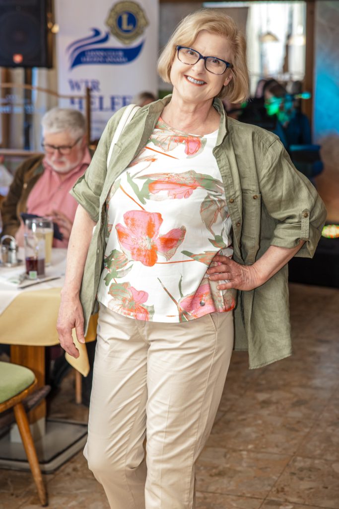 Eine lächelnde ältere Frau präsentiert bei einer Modenschau ein sommerlich-legeres Outfit. Sie trägt eine helle Hose, ein florales Shirt mit großen Blütenmustern in Korall- und Grüntönen sowie eine offene olivgrüne Leinenbluse. Mit einer Hand in der Hüfte posiert sie selbstbewusst im gemütlichen Veranstaltungsraum, während Gäste im Hintergrund an den Tischen sitzen und die Show verfolgen.