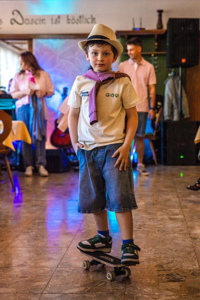 Ein kleiner Junge steht bei einer Modenschau selbstbewusst auf einem Skateboard mitten im Raum. Er trägt ein sommerliches Outfit mit Strohhut, weißem T-Shirt, Jeansshorts und einem locker über die Schultern gelegten violetten Pullover. Im Hintergrund sind weitere Teilnehmer und farbige Bühnenlichter zu sehen, während die gemütliche Atmosphäre des Veranstaltungsraums mit Holzdecken und warmem Licht erhalten bleibt.