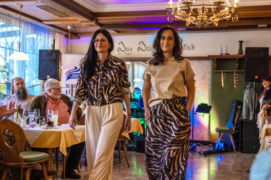 Zwei Frauen präsentieren bei einer Modenschau elegante Outfits mit auffälligem Zebra-Muster. Die Frau links trägt eine gemusterte Jacke zu einer hellen Hose, die Frau rechts einen passenden Rock mit schlichtem Oberteil. Beide gehen selbstbewusst durch einen gemütlichen Gastraum mit warmem Licht, während Gäste an den Tischen sitzen und die Show verfolgen. Im Hintergrund sorgen bunte Bühnenlichter und Musikequipment für eine festliche Atmosphäre.