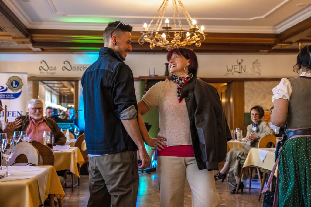 Ein junger Mann in dunkler Jacke und eine lächelnde Frau mit roten Haaren stehen bei einer Modenschau in einem gemütlichen Restaurant einander gegenüber und wirken ausgelassen und fröhlich. Die Frau trägt ein helles Outfit mit pinkem Oberteil und gemustertem Halstuch, dazu locker eine dunkle Jacke über der Schulter. Rundherum sitzen Gäste an gedeckten Tischen und beobachten die Szene, während warme Beleuchtung und farbige Bühnenlichter für eine stimmungsvolle Atmosphäre sorgen.