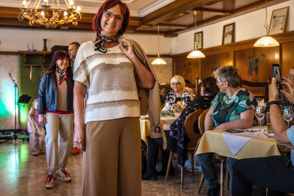 Eine lächelnde Frau mit roten Haaren präsentiert bei einer Modenschau ein leger-elegantes Outfit aus gestreiftem Strickoberteil und weiter beigefarbener Hose. Sie trägt ein gemustertes Halstuch und hat eine helle Jacke locker über die Schulter gelegt. Im Hintergrund läuft eine weitere Teilnehmerin über den Laufsteg, während die Gäste an gedeckten Tischen sitzen und die Veranstaltung verfolgen. Die warme Beleuchtung und Holzvertäfelung verleihen dem Raum eine gemütliche Atmosphäre.