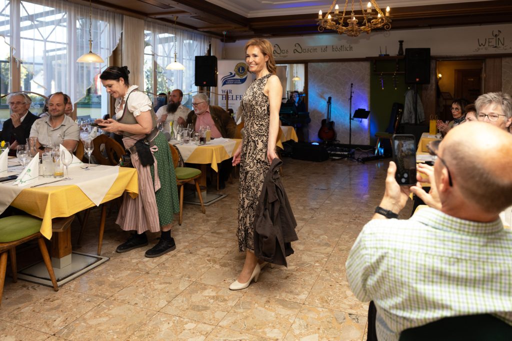 Eine Frau präsentiert bei einer Modenschau ein elegantes, ärmelloses Kleid mit floralem Muster in Schwarz und Beige. Sie läuft lächelnd durch einen festlich dekorierten Saal und hält eine dunkle Jacke locker in der Hand. Die Gäste sitzen an gedeckten Tischen und beobachten die Show, während im Vordergrund ein Besucher die Szene mit seinem Smartphone fotografiert. Warme Beleuchtung und Bühnenlicht schaffen eine stilvolle und gemütliche Atmosphäre.