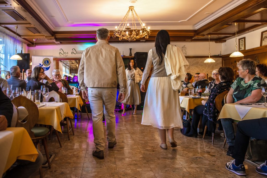 Ein Paar läuft bei einer Modenschau durch einen festlich beleuchteten Saal. Der Mann trägt ein helles Freizeitoutfit mit beiger Jacke und heller Hose, die Frau ein elegantes weißes Kleid mit grauer Weste und einem locker über die Schulter gelegten Cardigan. Die Gäste sitzen links und rechts an gedeckten Tischen und verfolgen aufmerksam die Präsentation. Im Hintergrund sorgt buntes Bühnenlicht für eine stimmungsvolle Atmosphäre.
