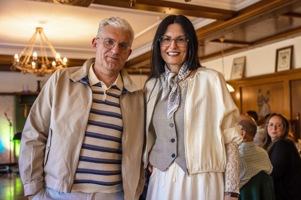 Ein stilvoll gekleidetes Paar posiert bei einer Modenschau in einem gemütlichen Veranstaltungssaal. Der Mann trägt eine helle Jacke über einem gestreiften Poloshirt in Beige- und Blautönen sowie eine helle Hose. Die Frau kombiniert eine weiße Hose mit einer grauen Weste, einer Spitzenbluse und einem gemusterten Halstuch. Beide lächeln in die Kamera, während im Hintergrund Gäste an Tischen sitzen und warmes Licht für eine elegante Atmosphäre sorgt.