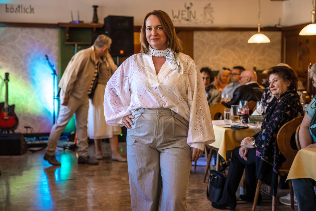 Eine Frau präsentiert bei einer Modenschau ein elegantes Outfit in hellen Naturtönen. Sie trägt eine weiße Bluse mit weitem Ärmel und feinem Muster, kombiniert mit einer hellbeigen Stoffhose und einem gemusterten Halstuch. Mit einer Hand in der Hüfte läuft sie selbstbewusst durch den Saal, während die Gäste an den Tischen zusehen. Im Hintergrund sorgen farbige Lichteffekte und eine Bühne mit Gitarre für eine stimmungsvolle Atmosphäre.