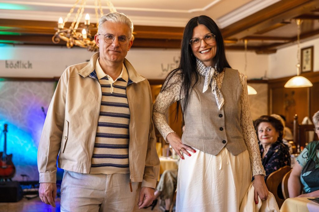 Ein Mann und eine Frau posieren gemeinsam bei einer Modenschau in einem stilvollen Veranstaltungssaal. Der Mann trägt eine helle Jacke über einem gestreiften Poloshirt mit passender heller Hose. Die Frau kombiniert ein cremefarbenes Kleid mit einer beigen Weste, Spitzenärmeln und einem gemusterten Halstuch. Beide lächeln in die Kamera, während im Hintergrund Gäste an gedeckten Tischen sitzen und die warme Beleuchtung für eine elegante Atmosphäre sorgt.