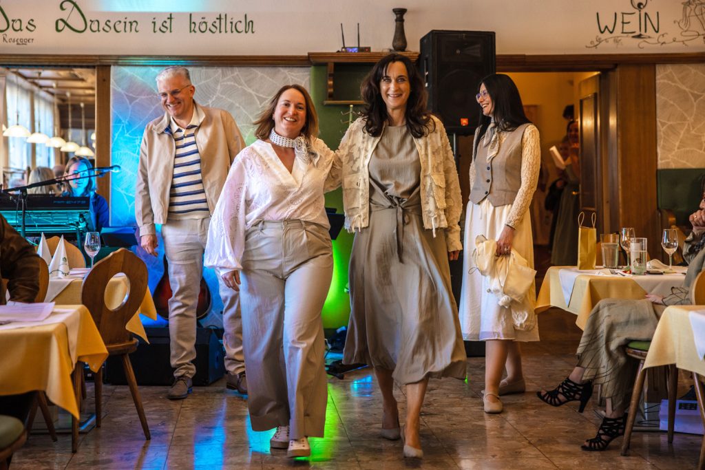 Vier Personen präsentieren gemeinsam elegante Outfits bei einer Modenschau in einem gemütlichen Veranstaltungssaal. Zwei Frauen laufen lächelnd vorneweg – eine in einer hellen Bluse mit weiter beiger Hose und die andere in einem fließenden beigen Kleid mit strukturierter Jacke. Dahinter folgen ein Mann in heller Freizeitmode und eine Frau in einem cremefarbenen Kleid mit Weste und Halstuch. Farbige Lichtakzente, eine Gitarre im Hintergrund und applaudierende Gäste schaffen eine warme und stilvolle Atmosphäre.