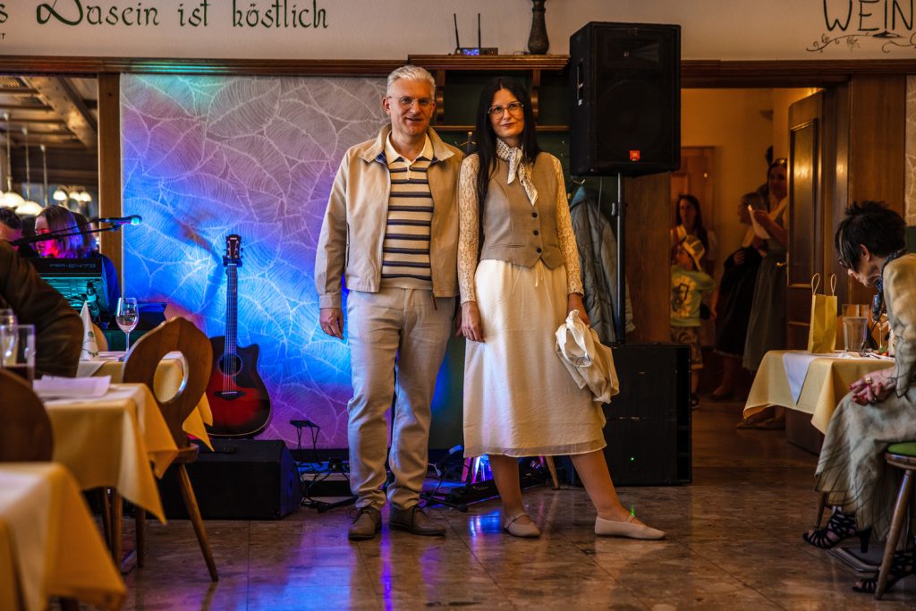 Ein Mann und eine Frau präsentieren gemeinsam elegante Outfits bei einer Modenschau in einem gemütlichen Veranstaltungssaal. Der Mann trägt eine helle Jacke über einem gestreiften Poloshirt mit passender heller Hose. Die Frau kombiniert ein cremefarbenes Kleid mit einer beigen Weste, Spitzenärmeln und einem gemusterten Halstuch. Im Hintergrund sorgen farbige Lichtakzente, eine Gitarre und die aufmerksam sitzenden Gäste für eine stilvolle Atmosphäre.