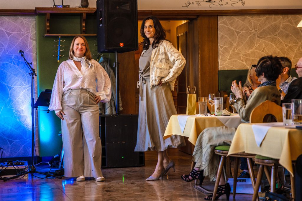 Zwei Frauen präsentieren elegante Outfits bei einer Modenschau in einem stilvollen Veranstaltungssaal. Links trägt eine Frau eine helle Bluse mit weitem Schnitt, kombiniert mit einer beigen Hose und einem gemusterten Halstuch. Rechts steht eine Frau in einem fließenden beigen Kleid mit gemusterter Jacke und hohen Schuhen. Die Gäste sitzen an gedeckten Tischen und beobachten die Präsentation aufmerksam, während farbige Lichtakzente für eine elegante Atmosphäre sorgen.