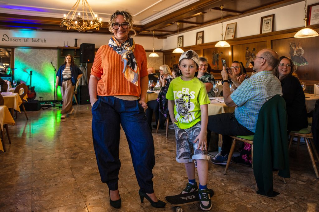 Eine Frau und ein Kind präsentieren gemeinsam Outfits bei einer Modenschau in einem gemütlichen Saal. Die Frau trägt einen orangefarbenen Pullover, einen gemusterten Schal, dunkle weite Hosen und schwarze Pumps. Neben ihr steht ein Junge in einem leuchtend grünen T-Shirt, gemusterten Shorts und einer Kappe rückwärts auf dem Kopf, mit einem Skateboard unter den Füßen. Im Hintergrund sitzen Gäste an gedeckten Tischen und beobachten die Szene aufmerksam.
