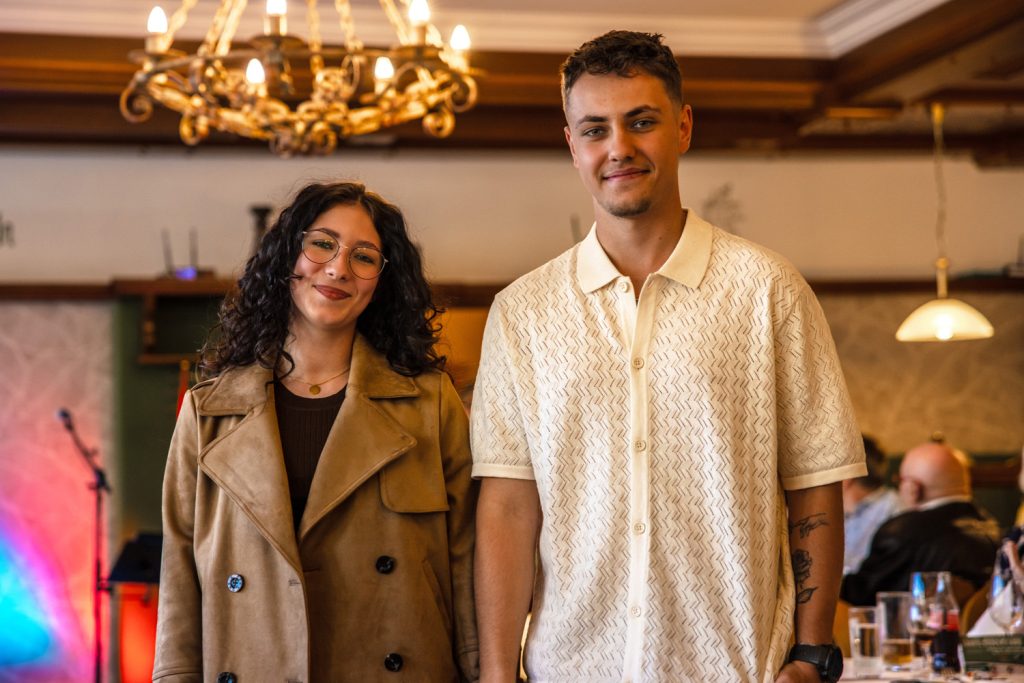 Zwei junge Erwachsene posieren gemeinsam bei einer Modenschau in einem gemütlichen Veranstaltungssaal. Die Frau trägt einen beigen Mantel über einem dunklen Oberteil und lächelt in die Kamera, während der Mann ein helles gemustertes Kurzarmhemd kombiniert mit einer beigen Hose trägt. Warmes Licht, ein Kronleuchter und farbige Bühneneffekte schaffen eine elegante und freundliche Atmosphäre.