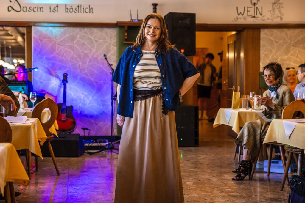 Eine Frau steht bei einer Modenschau selbstbewusst im Mittelpunkt des Laufstegs und präsentiert ein elegantes Outfit in natürlichen Beige- und Blautönen. Sie trägt einen langen beigen Rock, ein gestreiftes Shirt und ein dunkles blaues Overshirt. Im Hintergrund sorgen farbige Lichteffekte, eine Gitarre und die gemütliche Atmosphäre des Restaurants für einen stilvollen Rahmen, während Gäste aufmerksam zuschauen.