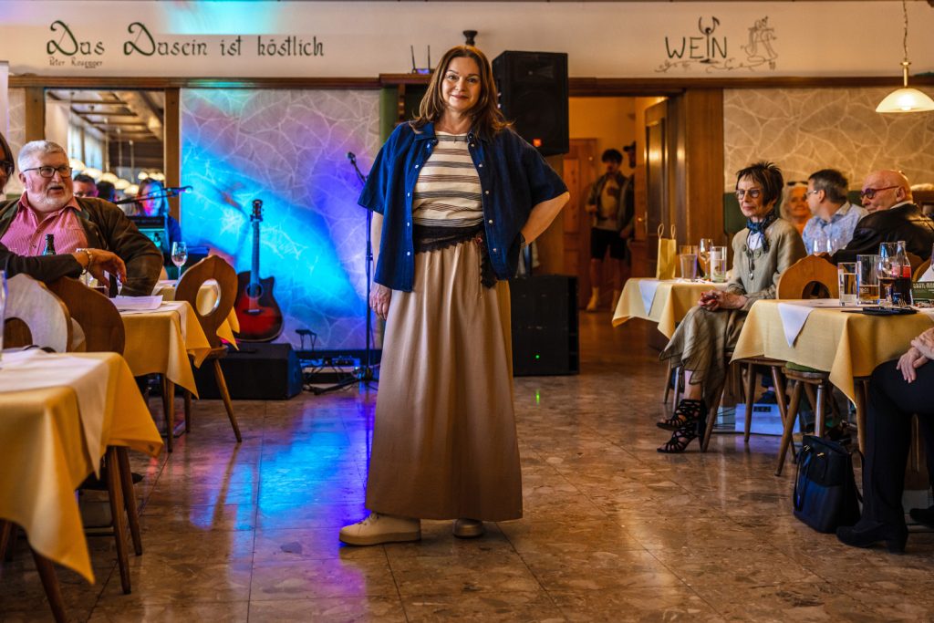 Eine Frau präsentiert bei einer Modenschau ein stilvolles Outfit in natürlichen Farben. Sie trägt einen langen beigen Rock, ein gestreiftes Oberteil und ein dunkles Overshirt, kombiniert mit hellen Sneakern. Die warme Atmosphäre des Saals mit bunten Lichtakzenten, einer Gitarre im Hintergrund und sitzenden Gästen verleiht dem Bild einen eleganten und zugleich gemütlichen Charakter.