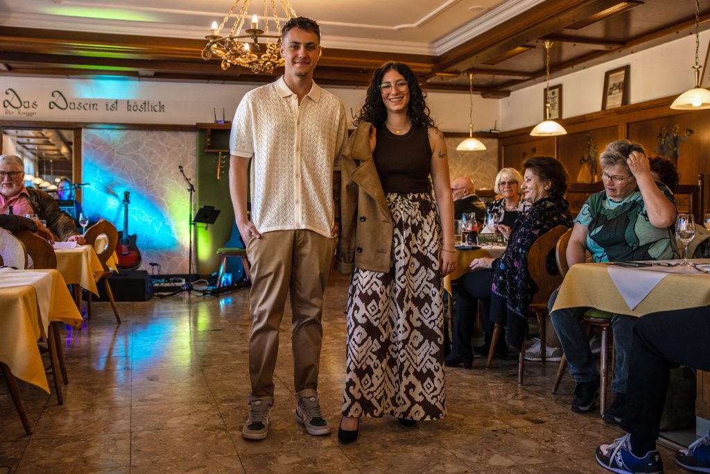 Zwei junge Models präsentieren elegante Freizeitmode auf einem Laufsteg in einem gemütlichen Veranstaltungssaal. Der Mann trägt ein helles gemustertes Kurzarmhemd mit brauner Hose und Sneakern, die Frau kombiniert ein dunkles Top mit einer gemusterten weiten Hose und einer lässig übergeworfenen Jacke. Die Gäste sitzen an den Tischen und verfolgen aufmerksam die stilvolle Modenschau.