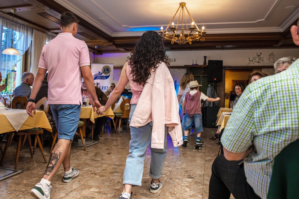 Zwei junge Models gehen Hand in Hand über den Laufsteg einer Modenschau in einem Restaurant. Der Mann trägt ein rosafarbenes Polo mit blauen Shorts, die Frau kombiniert eine helle Jeans mit rosafarbenem Oberteil und lässiger Jacke über der Schulter. Im Hintergrund sorgen Gäste, warme Beleuchtung und weitere Models für eine lockere und moderne Atmosphäre.
