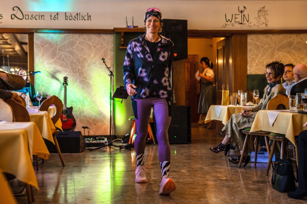 Eine Frau präsentiert sportliche Outdoor-Mode auf einem Laufsteg in einem gemütlichen Veranstaltungssaal. Sie trägt violette Leggings, eine dunkle gemusterte Jacke, Sportschuhe sowie Stirnband und Sonnenbrille auf dem Kopf. Im Hintergrund sorgen bunte Lichteffekte und eine Gitarre für Event-Stimmung, während die Gäste an den Tischen die Modenschau beobachten.