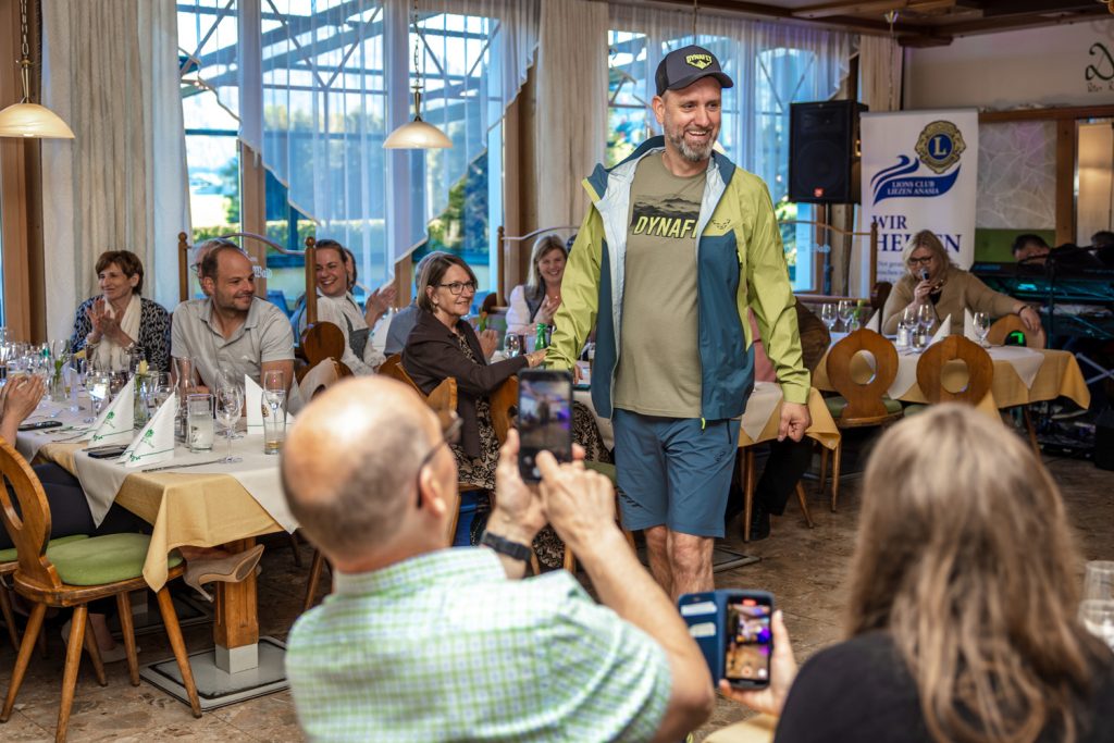 Ein Mann präsentiert sportliche Dynafit-Outdoorbekleidung bei einer Modenschau in einem Restaurant. Er trägt eine grün-blaue Regenjacke, passendes Shirt, Shorts und eine Kappe. Die Gäste sitzen links und rechts an den Tischen, applaudieren und filmen den Laufsteg mit ihren Smartphones. Die Stimmung wirkt locker, modern und sportlich.