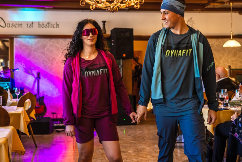 Zwei junge Models präsentieren moderne Dynafit-Sportmode auf einem Laufsteg in einem gemütlichen Veranstaltungssaal. Die Frau trägt ein sportliches Outfit in kräftigem Pink mit passender Sonnenbrille, der Mann kombiniert ein blaues Dynafit-Shirt mit lockerer Outdoor-Jacke und Stirnband. Beide gehen Hand in Hand und sorgen mit ihren sportlichen Looks für eine dynamische Atmosphäre.