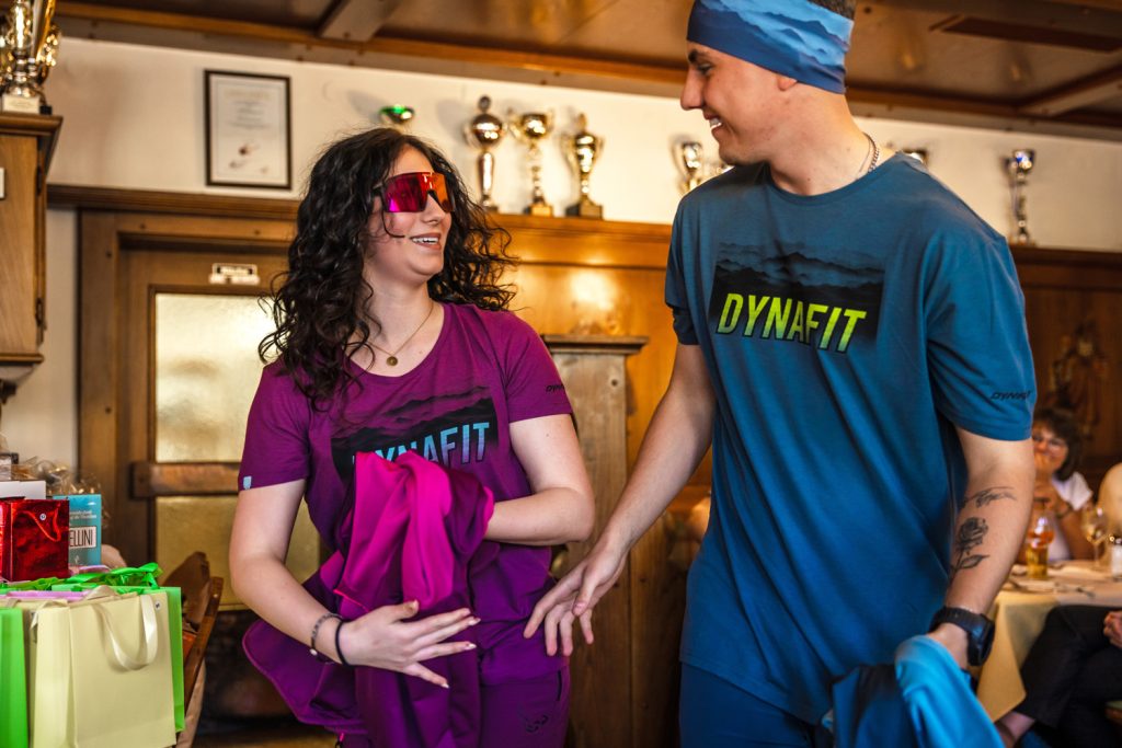 Zwei junge Models präsentieren sportliche Outdoor-Mode bei einer Modenschau in gemütlicher Atmosphäre. Die Frau trägt ein pinkes Dynafit-Shirt, passende Sportbekleidung und eine auffällige verspiegelte Sonnenbrille. Der Mann kombiniert ein blaues Dynafit-Shirt mit Stirnband und sportlichem Outfit. Beide lachen sich an und wirken locker und dynamisch, während im Hintergrund Pokale und Gäste zu sehen sind.
