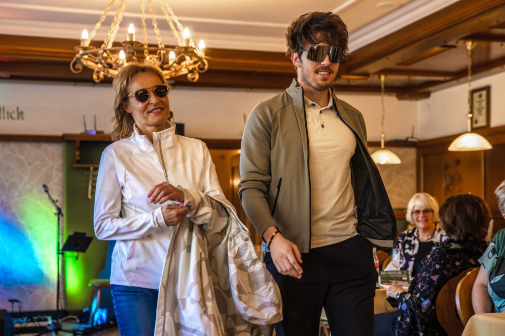Zwei Models präsentieren sportlich-elegante Freizeitmode bei einer Modenschau in einem stilvollen Veranstaltungssaal. Die Frau trägt eine helle Funktionsjacke mit Jeans und Sonnenbrille und hält einen passenden gemusterten Mantel in der Hand. Der Mann kombiniert ein helles Poloshirt mit leichter Jacke, dunkler Hose und auffälliger Sonnenbrille. Warme Beleuchtung, Holzelemente und applaudierende Gäste schaffen eine moderne und entspannte Atmosphäre.