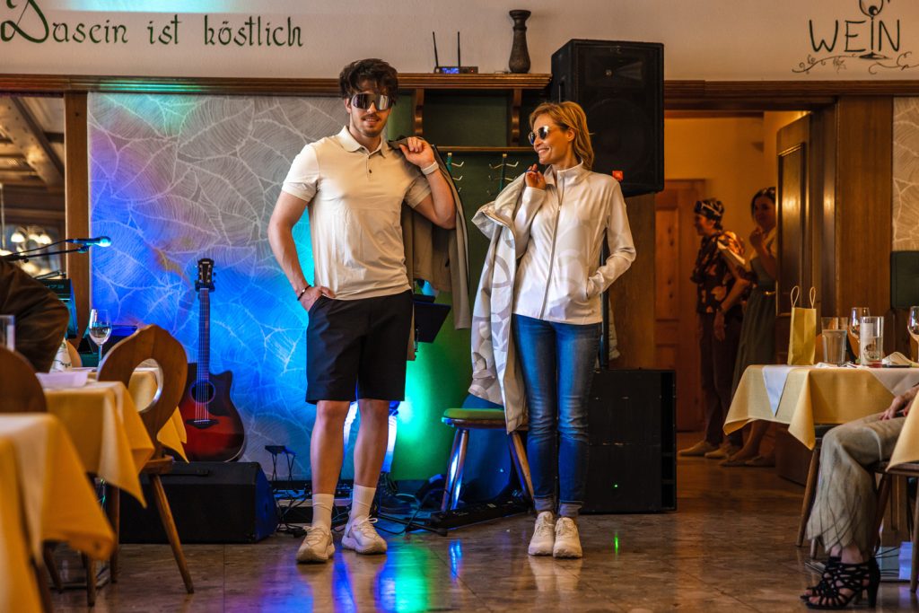 Zwei Models präsentieren moderne Freizeitmode bei einer Modenschau in einem gemütlichen Veranstaltungssaal. Der Mann trägt ein helles Poloshirt mit dunklen Shorts und Sonnenbrille, die Frau eine helle gemusterte Jacke mit Jeans und Sonnenbrille. Beide halten locker ihre Jacken über der Schulter, während bunte Lichtakzente und sitzende Gäste die stilvolle Atmosphäre ergänzen.