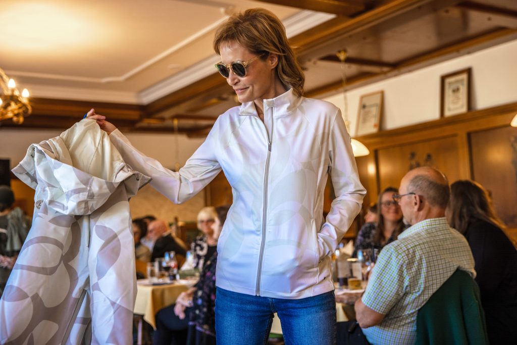 Eine Frau präsentiert bei einer Modenschau ein sportliches Freizeitoutfit mit heller gemusterter Jacke, Jeans und Sonnenbrille. In der Hand hält sie einen passenden Mantel mit dezentem Muster. Die Szene spielt in einem warm beleuchteten Veranstaltungssaal mit Holzwänden und sitzenden Gästen, die die Modenschau verfolgen.