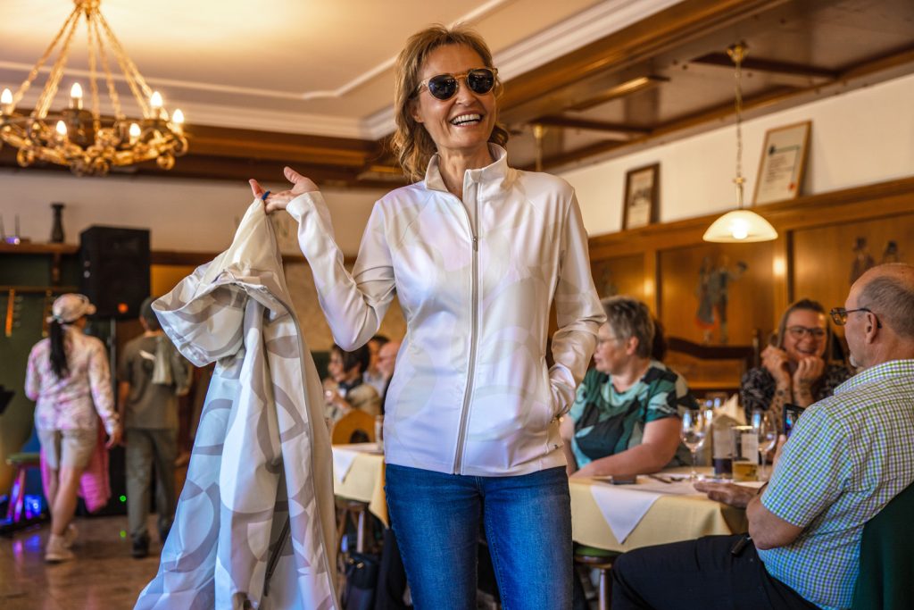 Eine lächelnde Frau präsentiert bei einer Modenschau ein sportlich-elegantes Freizeitoutfit mit heller Jacke, Jeans und Sonnenbrille. In der Hand hält sie einen gemusterten Mantel. Die Szene spielt in einem gemütlichen Veranstaltungssaal mit Holzelementen, Kronleuchter und sitzenden Gästen, die die Modenschau aufmerksam verfolgen.