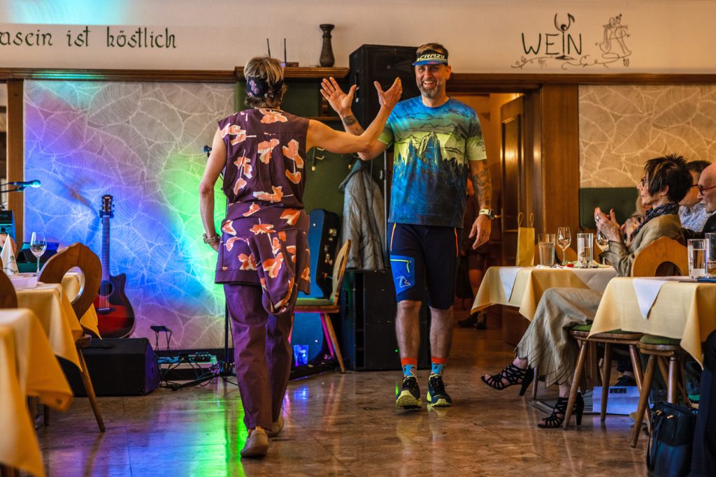 Bei einer Modenschau in einem rustikalen Veranstaltungssaal begrüßen sich zwei Models mit einem High-Five auf dem Laufsteg. Die Frau trägt ein violett gemustertes Freizeit-Outfit mit Stirnband, der Mann ein farbenfrohes Sportshirt mit blauen Shorts und Stirnband. Bunte Lichtakzente, eine Gitarre und applaudierende Gäste schaffen eine lockere und lebendige Atmosphäre.
