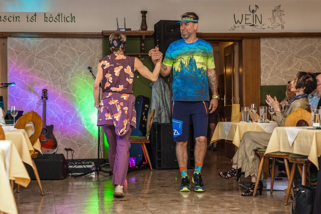 Zwei Models präsentieren sportliche Outdoor-Mode bei einer Modenschau in einem traditionellen Veranstaltungssaal. Eine Frau in einem violett gemusterten Freizeit-Outfit und ein Mann in einem bunten Funktionsshirt mit blauen Shorts geben sich die Hand auf dem Laufsteg. Im Hintergrund sorgen farbige Lichteffekte, eine Gitarre und applaudierende Gäste für eine lebendige Atmosphäre.